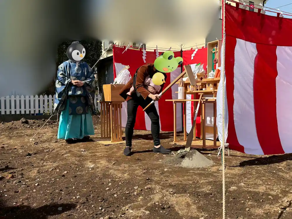 青空の下、地鎮祭で「鍬入れ（くわいれ）」の儀式を行う家族の様子。土地活用会社の元営業課長である筆者の夫が、娘を抱っこしながら力強く砂の山に鍬を入れ、家族全員で新しい家づくりへの決意を固めている。背景はプライバシー保護のため加工されているが、清々しい快晴の中での特別な一日の臨場感を表現している。